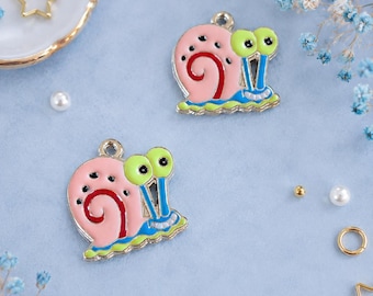 Op de afbeelding: Twee roze slakbedels met groene ogen, rode wervelingen en blauwe en gele accenten. Deze emaille bedels hebben een zilverkleurige lus aan de bovenkant om aan sieraden of knutselwerk te bevestigen. Op een lichtblauwe ondergrond.