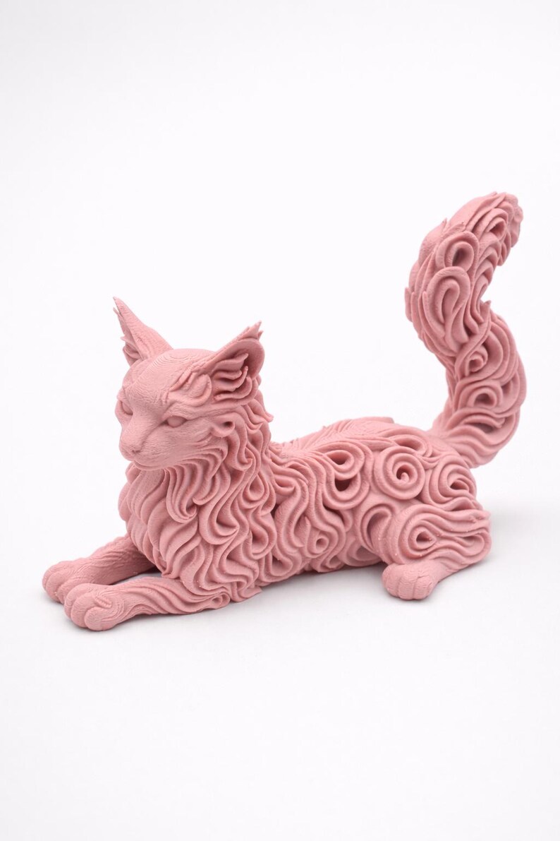 Op de afbeelding: Een roze kattenbeeld in rustpositie. De vacht van de kat heeft een gedetailleerd wervelend patroon. De sculptuur heeft puntige oren en een lange, gekrulde staart. De kat ligt op een witte achtergrond.