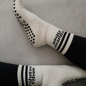 Könnte beinhalten: Weiße Pilates-Socken mit schwarzen Grip-Punkten auf den Sohlen und dem Text "Pilates Vibes" in Schwarz. Die Socken haben schwarze Streifen am oberen Rand.