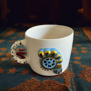 May include: White ceramic mug with a blue and gold decorative design on the front and handle. The handle features a polka dot pattern with red accents. The mug is set against a patterned teal and orange background.