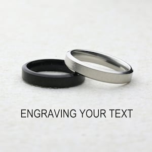 Peut inclure: Deux anneaux simples, l'un noir mat et l'autre argent brossé, posés côte à côte. Le texte ENGRAVING YOUR TEXT est imprimé en lettres capitales noires sous les anneaux, indiquant la possibilité de personnalisation sur ces anneaux en métal.