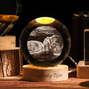 Può includere: Una sfera di cristallo con un'immagine ecografica di un bambino, illuminata da una luce calda. La sfera poggia su una base di legno con l'iscrizione "To my baby Rose". Altri oggetti di cristallo sono sullo sfondo.