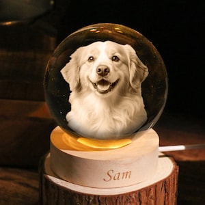 May include: A clear glass sphere with a smiling dog portrait, resting on a wooden base with the name "Sam" engraved. The base sits on a rustic wood slice, illuminated by a warm light. The dog's image is in black and white.