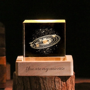 May include: A glowing, clear glass cube with a detailed solar system design. The cube sits on a wooden base engraved with the words "You are my universe". The base rests on a rustic wooden stand, creating a warm, inviting aesthetic.