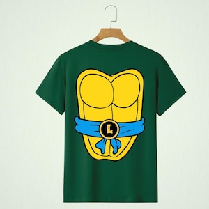May include: Green t-shirt featuring a yellow turtle shell design with a blue belt and a gold buckle with the letter "L". The shirt is hanging on a wooden hanger.