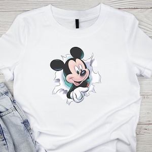 May include: White t-shirt featuring a graphic of Mickey Mouse bursting through a torn hole. The cartoon character is in black, red, and white, with a friendly expression. The shirt is made of a soft material and is suitable for casual wear.