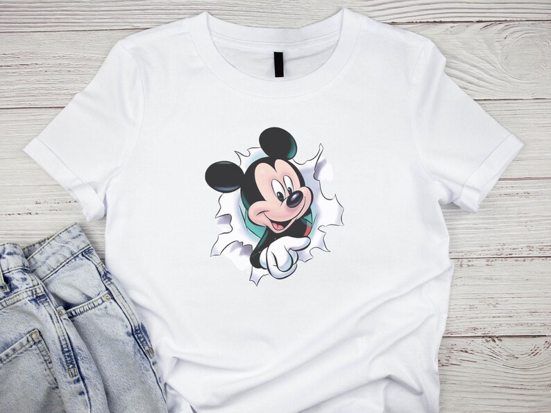 May include: White t-shirt featuring a graphic of Mickey Mouse bursting through a torn hole. The cartoon character is in black, red, and white, with a friendly expression. The shirt is made of a soft material and is suitable for casual wear.
