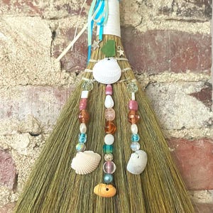May include: A decorative broom with a natural straw brush head. The handle is wrapped in white paper and adorned with seashells, beads, and ribbons in shades of blue, pink, and orange. Small star and sea glass accents add to the coastal theme.