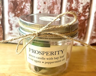 May include: A clear glass jar candle labeled "PROSPERITY" with a green candle, bay leaf, moss agate, and peppermint oil. The jar is tied with twine and has a silver lid. The background is a brick wall.