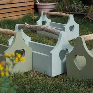 May include: A collection of wooden garden boxes in shades of green and blue-green. Each box features a heart-shaped cutout and a natural wood handle. The boxes are on grass.