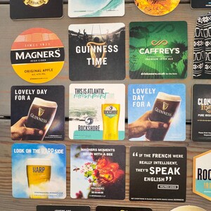 Op de afbeelding: Verzameling bier- en cideronderzetters met verschillende merken zoals Guinness, Magners en Rockshore. De onderzetters tonen logo's, slogans en afbeeldingen gerelateerd aan de dranken, met een houten achtergrond.