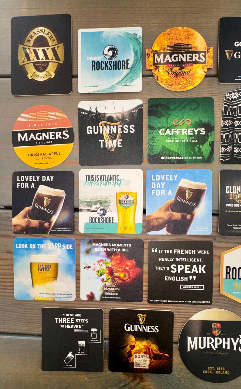 Op de afbeelding: Verzameling bier- en cideronderzetters met verschillende merken zoals Guinness, Magners en Rockshore. De onderzetters tonen logo's, slogans en afbeeldingen gerelateerd aan de dranken, met een houten achtergrond.