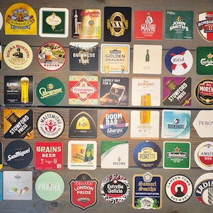 Op de afbeelding: Een verzameling bierviltjes met verschillende merklogo's en ontwerpen. De viltjes zijn rond, vierkant en rechthoekig en hebben een verscheidenheid aan kleuren en tekst. Merken zijn Guinness, Heineken en Budvar.