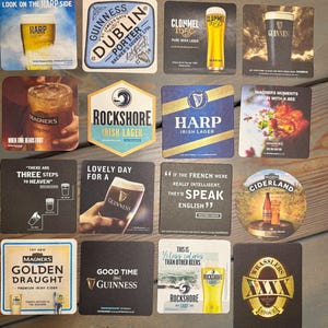 Puede incluir: Un conjunto de posavasos de cerveza y sidra con marcas como Guinness, Harp y Magners. Los posavasos muestran im&aacute;genes de bebidas, logotipos y texto, con varias formas y dise&ntilde;os. Los posavasos est&aacute;n sobre una superficie de madera.