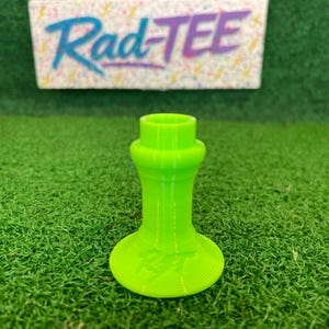 Può includere: Un tee da golf verde neon con base svasata e parte superiore cilindrica. Il tee è su uno sfondo di erba verde, con un cartello che recita "Rad-Tee" in un font colorato e stilizzato.