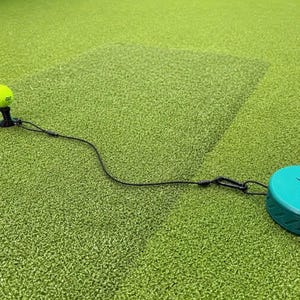May include: A neon yellow golf ball sits on a black tee, connected by a thin black cord to a round teal anchor disc. The disc features the letters RT embossed on its top surface. The entire assembly is designed for golf practice and training.