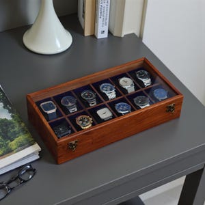 May include: A wooden watch display case with a glass top, showcasing twelve watches. The case is rectangular with a rich brown finish and brass-colored clasps. Each watch rests on a dark blue velvet cushion, offering a stylish storage solution.