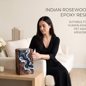 May include: A rectangular urn made of Indian rosewood and epoxy resin. The urn features a dark wood grain pattern with a blue and white wave design on the front. The text on the image reads "Suitable for: Human Ashes, Pet Ashes, Memories."