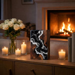 May include: A decorative wooden vase with a black and white resin design, displayed on a wooden cabinet. The vase is rectangular and stands near a bouquet of white roses in a glass vase and several lit candles. A fireplace is in the background.
