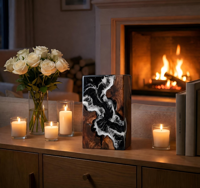 May include: A decorative wooden vase with a black and white resin design, displayed on a wooden cabinet. The vase is rectangular and stands near a bouquet of white roses in a glass vase and several lit candles. A fireplace is in the background.