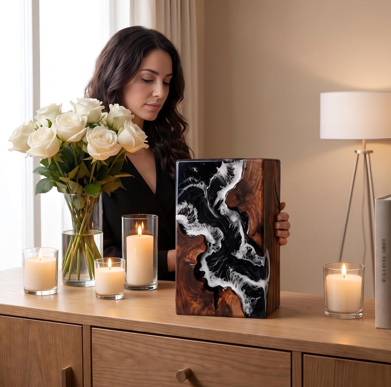 May include: A wooden decorative box with a black and white resin design. The box is rectangular and sits on a wooden surface with lit candles and a bouquet of white roses. A woman is holding the box.