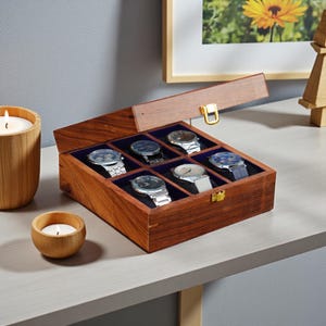 May include: A wooden watch box with a hinged lid, holding six wristwatches with various band styles. The box is made of dark wood with a blue velvet interior. A gold-colored clasp secures the lid.