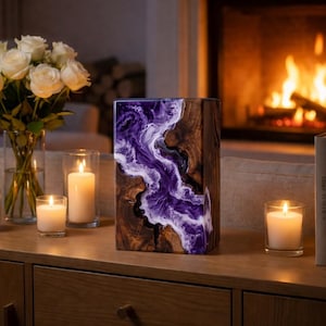 May include: A rectangular wooden art piece with a river of purple and white resin. The wood has a natural grain pattern. The piece is on a wooden surface with candles and a vase of white roses.