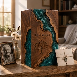 May include: A rectangular wooden cremation urn featuring a central vertical inlay of swirling teal resin. The front surface is engraved with the name David Morgan, the dates 1955-2023, and a detailed tree of life illustration.