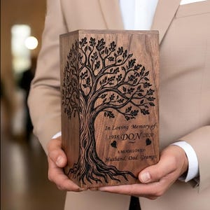 Pode incluir: Uma urna de madeira com um desenho de árvore da vida, segurada por uma pessoa. A urna tem o texto gravado "In Loving Memory of 1938 DON 2020 A MUCH LOVED Husband, Dad, Gramps". A urna é retangular e de madeira castanho claro.
