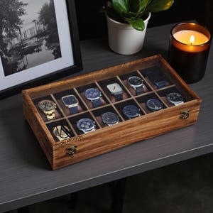 Puede incluir: Una caja de exhibición de relojes de madera con tapa de cristal transparente, que muestra una colección de varios relojes de pulsera. La caja es rectangular con un acabado de madera cálida y cierres de color latón. Los relojes tienen correas y esferas de diferentes colores.