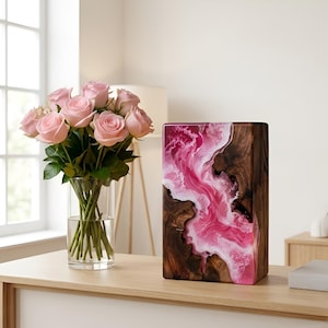 May include: A decorative wooden block with a vibrant pink and white resin design. The block is rectangular and stands upright on a wooden surface. A bouquet of pink roses in a clear glass vase is to the left.