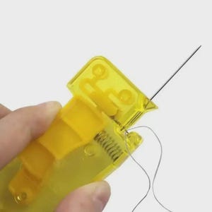 May include: A yellow plastic needle threader with a metal needle and gray thread. The threader has a hinged design with a spring mechanism. The needle is thin and sharp, designed for sewing. The threader is held in a hand.