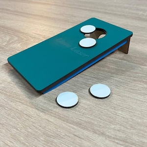 Op de afbeelding: Tealkleurig miniatuur cornhole-spel met drie witte beanbags bovenop en twee eronder. Het speelbord heeft een blauwe rand en wordt ondersteund door een bruine steun. De tekst "Erin Laser" is zichtbaar.