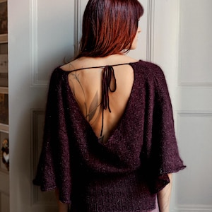 May include: A dark purple knit top with a deep V-shaped back opening and short sleeves. The top is tied at the back of the neck with a matching fabric tie. The model is wearing matching purple pants.