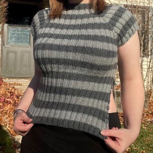 Puede incluir: Un top de punto a rayas grises y gris oscuro con mangas cortas y cuello alto. El top tiene una textura acanalada y una abertura lateral con botones. Una prenda elegante para cualquier guardarropa.