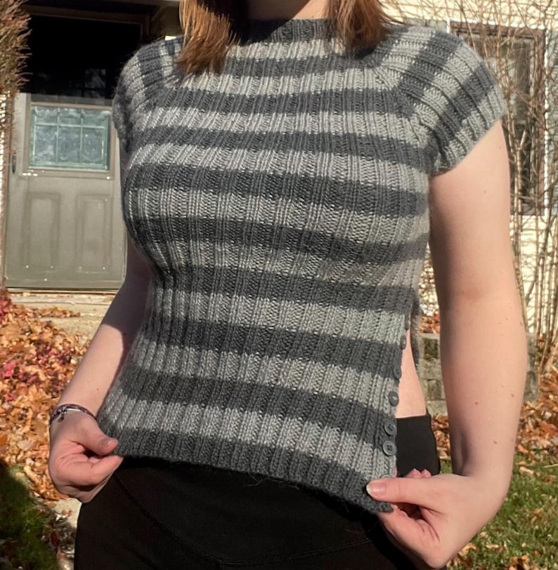 Puede incluir: Un top de punto a rayas grises y gris oscuro con mangas cortas y cuello alto. El top tiene una textura acanalada y una abertura lateral con botones. Una prenda elegante para cualquier guardarropa.
