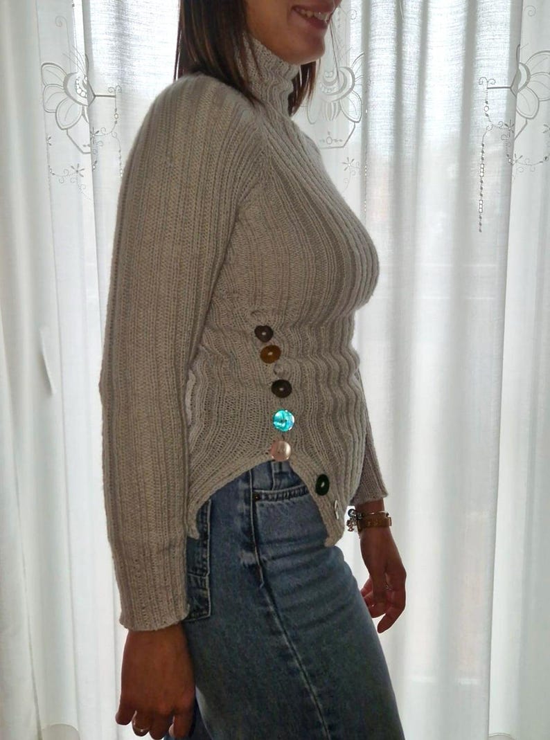 Puede incluir: Un jersey de cuello alto beige claro con canal&eacute; vertical y mangas largas. El jersey presenta botones decorativos de varios colores en el lateral. La persona lleva vaqueros azules.