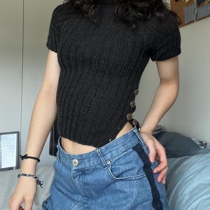 Puede incluir: Una parte superior de punto negra de manga corta con cuello alto y botones decorativos en el lateral. La parte superior se combina con unos vaqueros azules. Los vaqueros tienen una raya azul oscuro en el lateral.