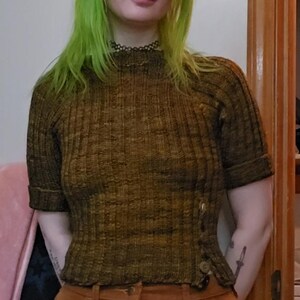 Puede incluir: Top de punto acanalado verde oliva de manga corta con botones laterales, combinado con pantalones de pana marrones. El conjunto es de tonos tierra y casual.