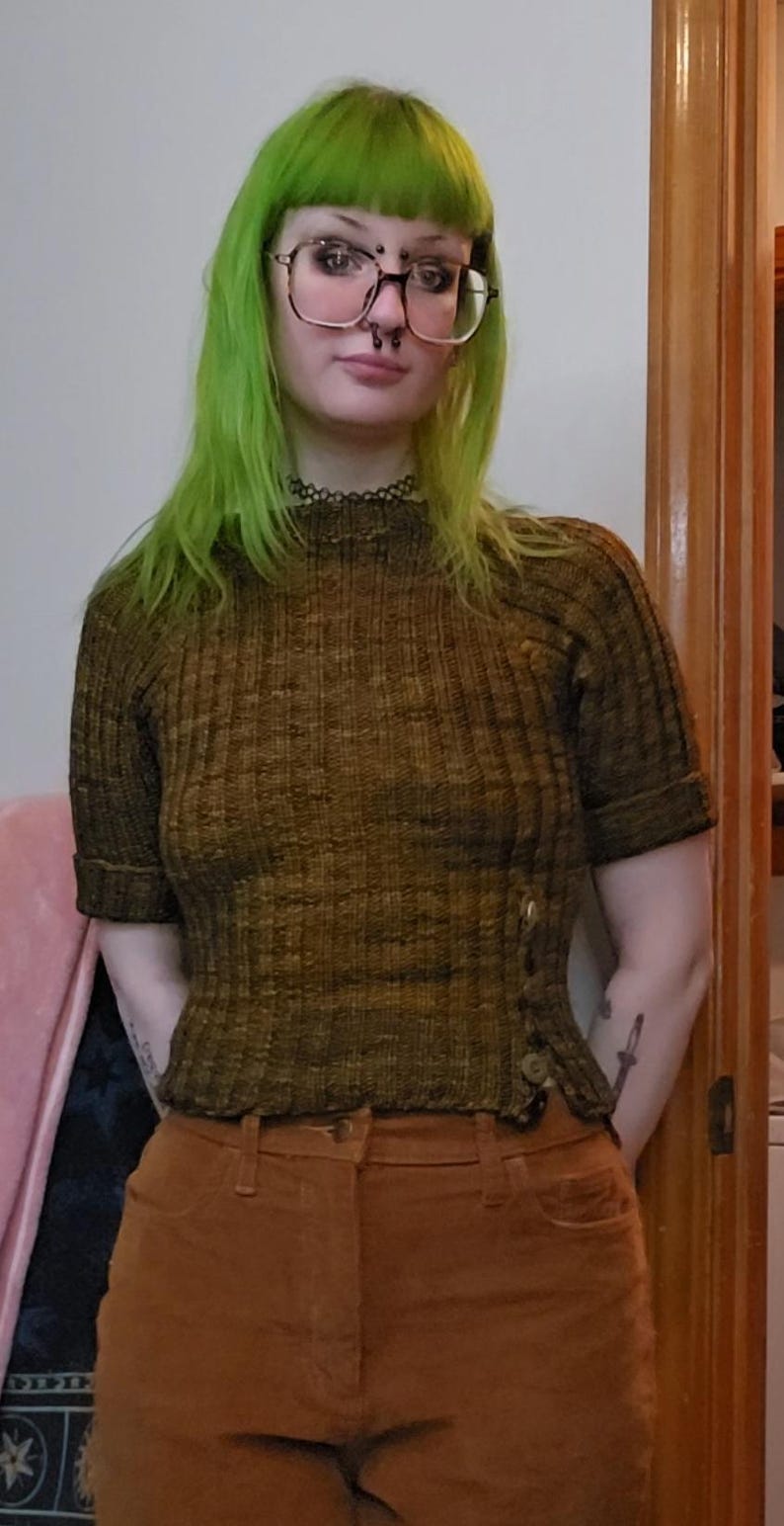 Puede incluir: Top de punto acanalado verde oliva de manga corta con botones laterales, combinado con pantalones de pana marrones. El conjunto es de tonos tierra y casual.
