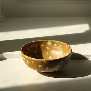 May include: A small, round ceramic bowl with a deep mustard-yellow glaze. The exterior and interior surfaces are decorated with irregular, off-white, hand-painted spots that vary in size and shape, creating a speckled pattern across the entire piece.