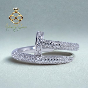 May include: A silver-colored ring with a nail-head design, encrusted with small, sparkling stones. The ring features a curved band and a vertical post, creating a unique and modern look. The ring is set against a light background.