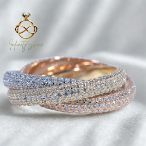 Puede incluir: Anillo de tres bandas con engaste de pavé. El anillo presenta bandas en plata, oro y oro rosa, todas incrustadas con pequeñas piedras brillantes. Las bandas están entrelazadas, creando un diseño único y elegante. El logo "Infinity Shine" está en la esquina superior izquierda.