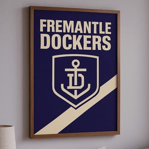 Puede incluir: Póster enmarcado con las palabras "FREMANTLE DOCKERS" en blanco sobre un logotipo del equipo. El logotipo es un ancla blanca dentro de un escudo, sobre un fondo azul marino con una franja diagonal beige. El marco es marrón.