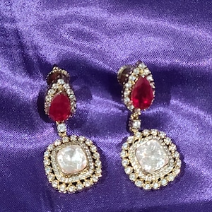 May include: A pair of gold-toned drop earrings. Each earring features a teardrop-shaped red gemstone at the top, connected to a square, off-white stone surrounded by small, clear crystals. The earrings are set against a purple background.