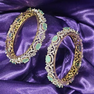 May include: Two circular gold-tone bangles featuring an intricate openwork metal design encrusted with small clear stones and accented with evenly spaced, oval-shaped light green cabochon stones set along the outer circumference of each piece.