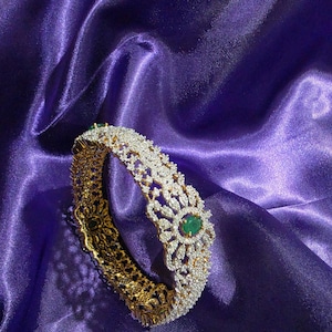May include: A gold-toned bangle bracelet featuring an intricate filigree design encrusted with numerous small clear stones. The center of the floral-inspired pattern is accented with a single oval-shaped green gemstone, creating a contrast.