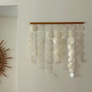 May include: A wall hanging featuring a horizontal wooden rod from which seven vertical strands of translucent, circular capiz shell discs are suspended. The discs vary in size and are arranged in cascading lengths, creating a layered effect.