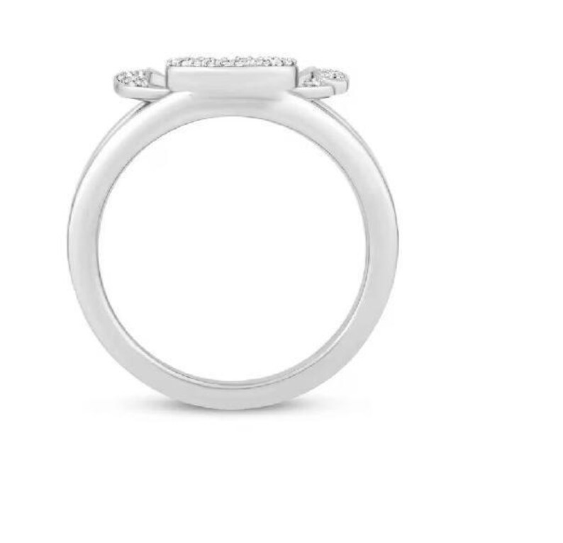 Op de afbeelding: Een zilveren ring met een rechthoekige middensteen en kleine accentstenen aan weerszijden. De ringband is glad en afgerond. De ring staat tegen een witte achtergrond.