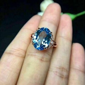 以下が含まれることがあります： 大きな楕円形のライトブルーの宝石をあしらったローズゴールドのリング。宝石は、より小さな透明なアクセントストーンで囲まれています。リングは、暗い背景の中で手に表示されています。
